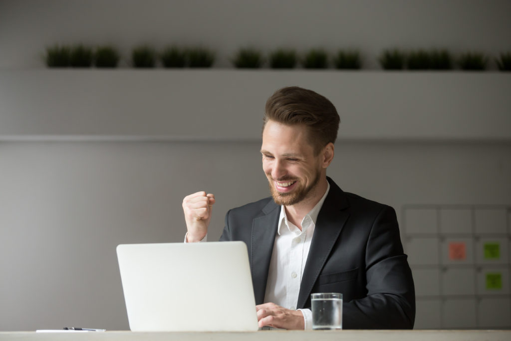 Man celebrating success in trading with yes gesture and looking at laptop.